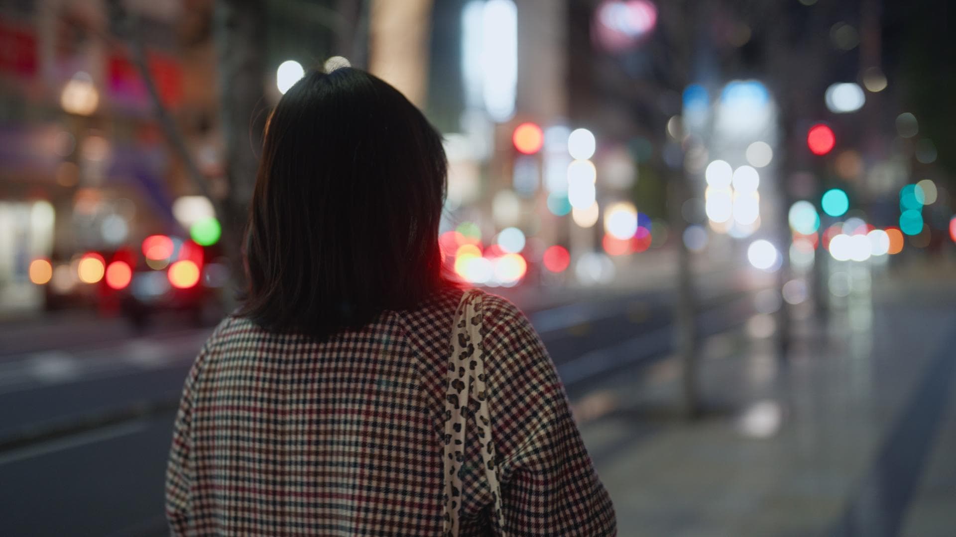 Eine Frau spaziert allein durch eine große, hellbeleuchtet Stadt. Sie kerht uns ihren Rücken zu und trägt einen karierten Herbstmantel.