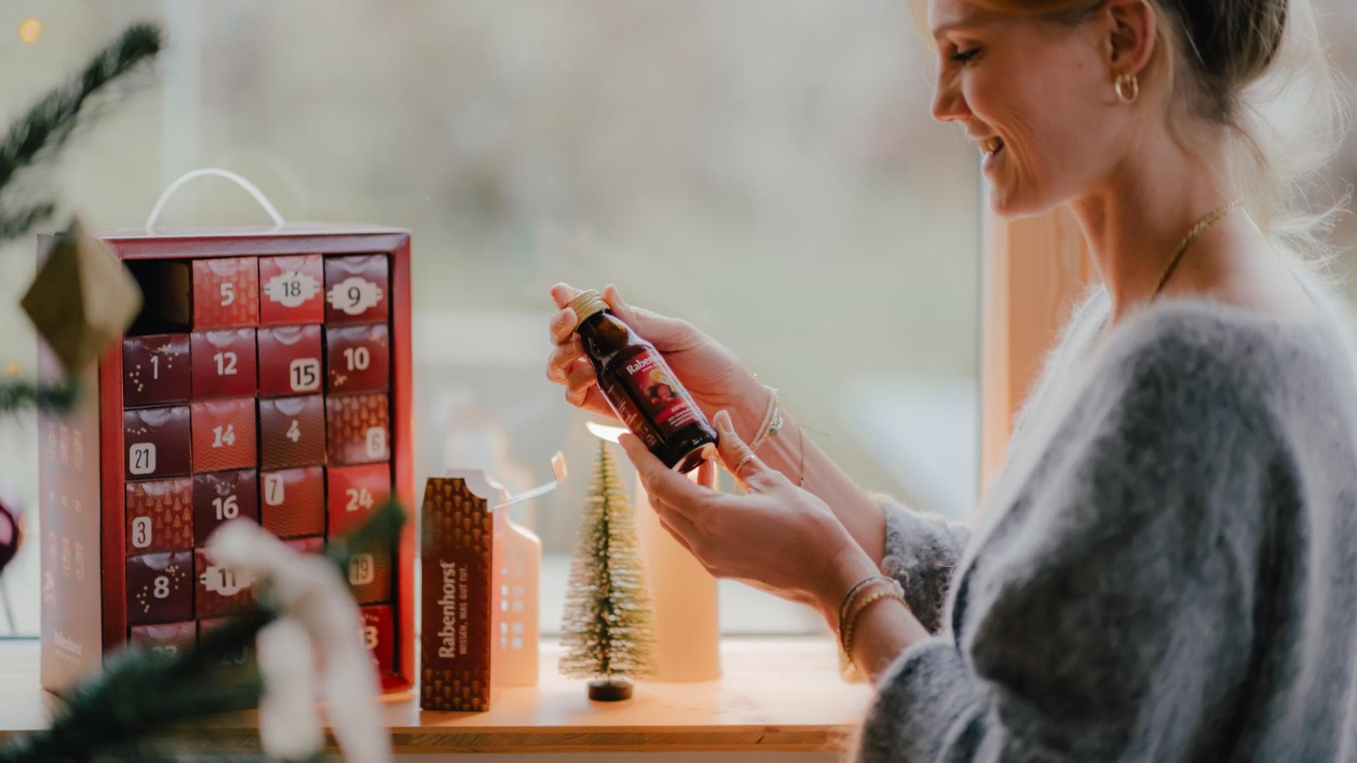 Eine Frau hat das erste Türchen ihres Rabenhorst-Adventskalenders geöffnet und hält freudig strahlend einen Rabenhorst-Mini-Saft in der Hand 