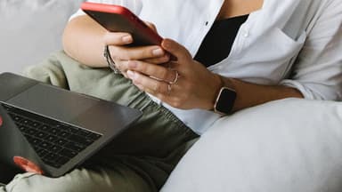 Eine Frau mit Laptop auf dem Schoß checkt ihr Smartphone. 