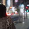 Eine Frau spaziert allein durch eine große, hellbeleuchtet Stadt. Sie kerht uns ihren Rücken zu und trägt einen karierten Herbstmantel.