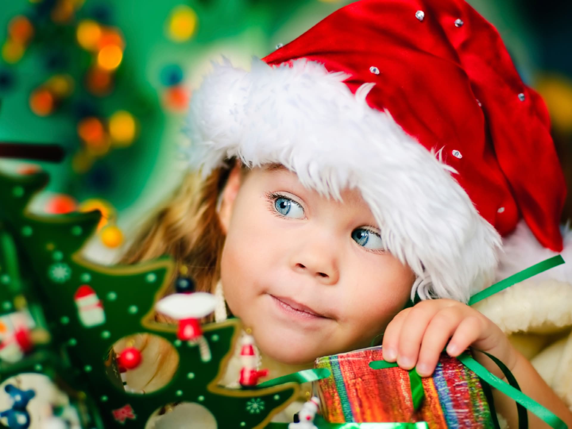 Kleines Mädchen mit Weihnachtsmütze lugt hinter dem Tannenbaum hervor